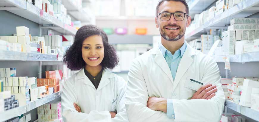 Two pharmacists stand behind the pharmacy counter with arms crossed, smiling.