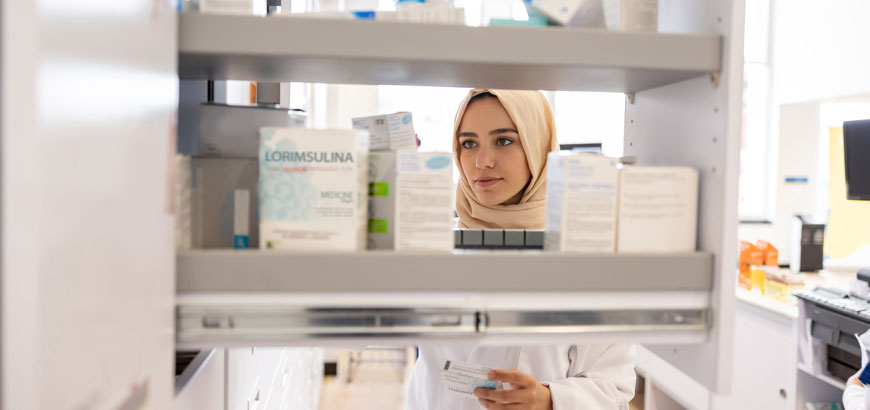 A female pharmacist wearing a hijab is seen from the other side of a medication shelf