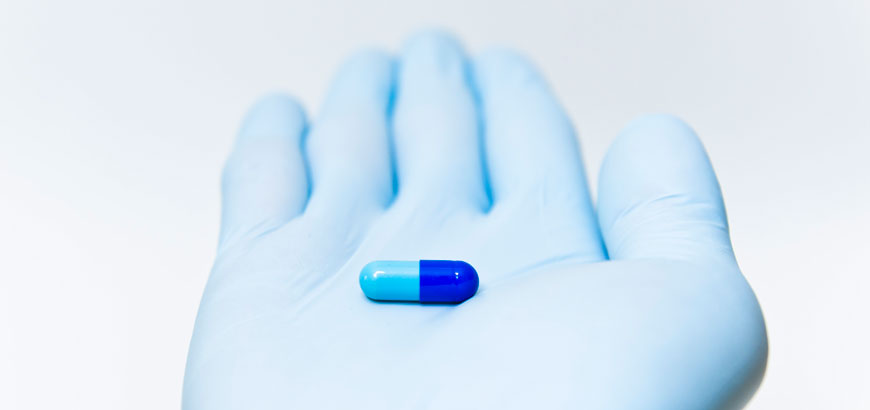 A hand in a blue medical glove holding a prescription pill.