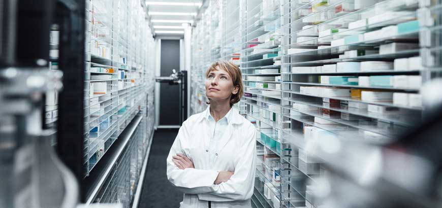 A pharmacist smiles and looks up at shelves full of drugs