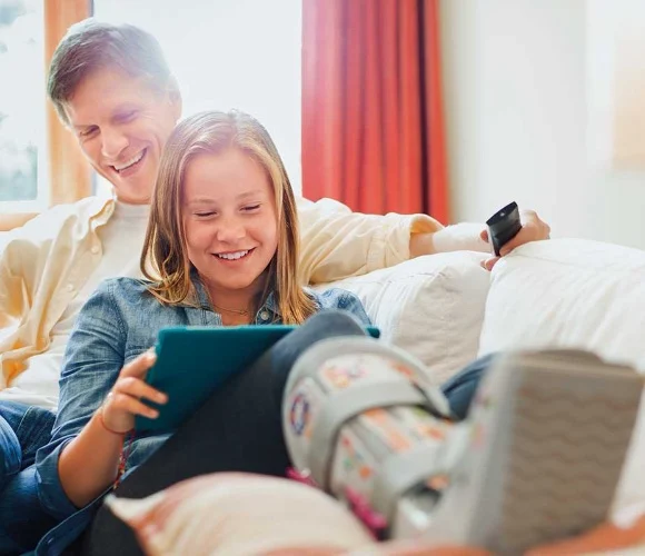 parent sitting on couch with child with cast on their leg both smiling looking at a tablet