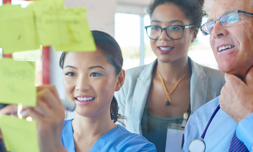 Three Healthcare workers stand together and make notes on post it's hung on a wall.