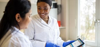 A pharmacist smiles at another pharmacist while holding a tablet