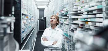 A pharmacist stands among the shelves of medications, looking upwards