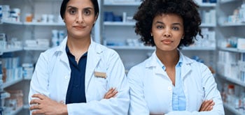 Two female pharmacists stand in the pharmacy, arms crossed, looking right into the camera