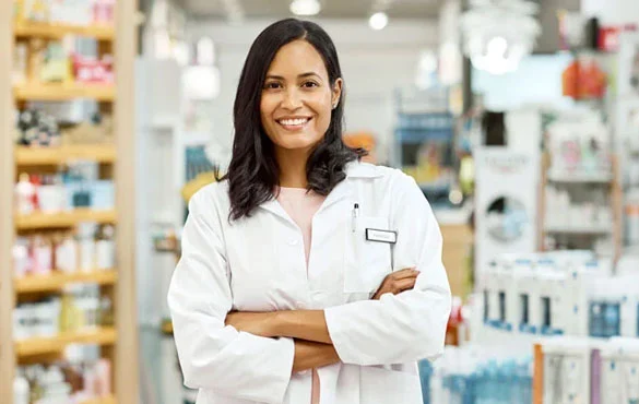 Pharmacist in white coat standing in a pharmacy with arms crossed.