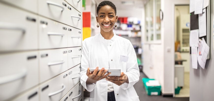 <span>Happy female pharmacist standing by a medicine shelf holding digital tablet.</span>