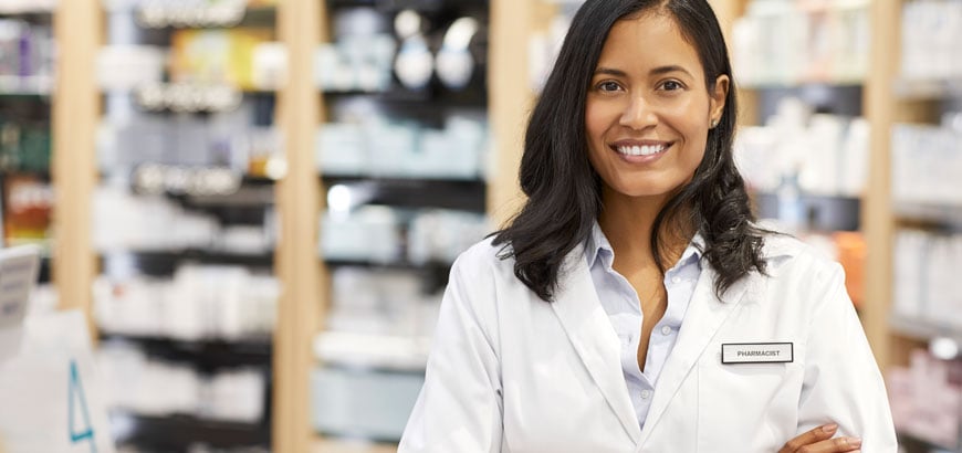 A female pharmacist stands with her arms crossed inside her pharmacy. She is wearing a white lab coat with a name tag that says &quot;pharmacist&quot; on it.