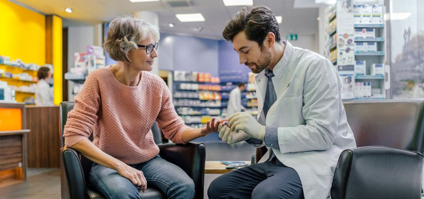 A pharmacist sits in a chair across from a patient and pricks her finger for a blood test