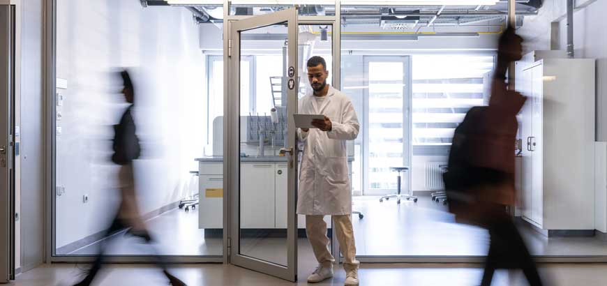 A doctor standing in a doorway checking a clipboard while other people walk quickly by him