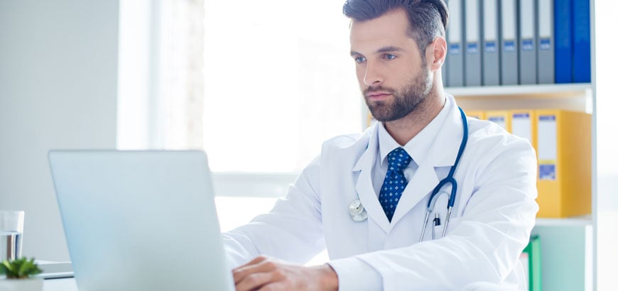 A male health care worker sits at a desk and types on a laptop computer.