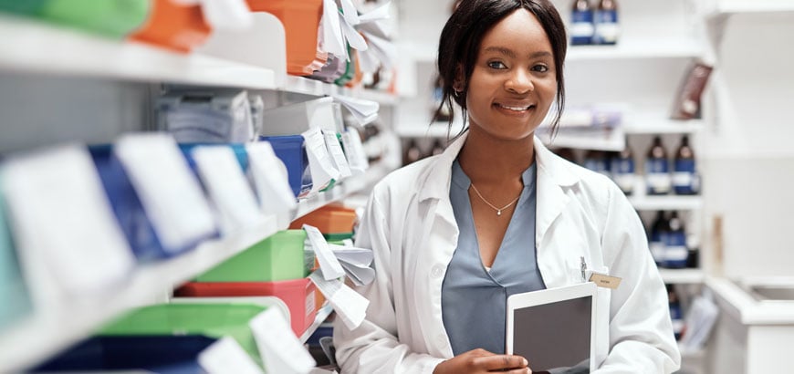 A pharmacist stands behind the counter holding an iPad in her arms