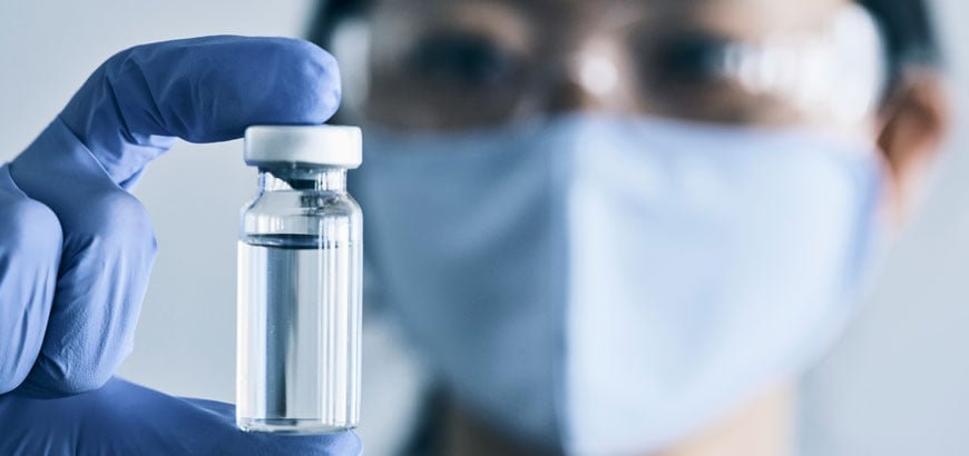 A close up on a medicine vial with a woman wearing safety glasses behind it.