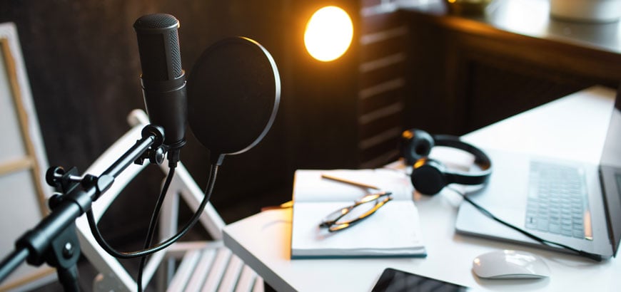 A close up of a desk with a microphone, tablet, paper notebook and laptop open on it.