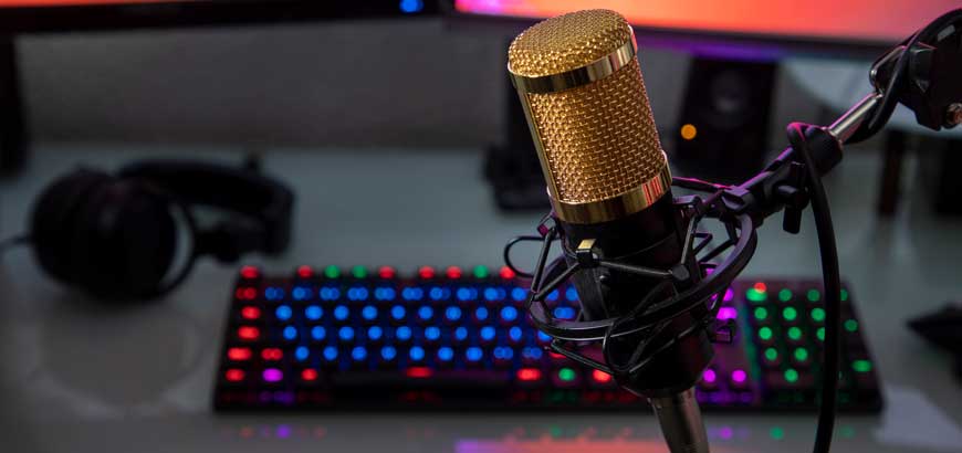 A recording microphone in front of two computer monitors and a lit up multcolored keyboard