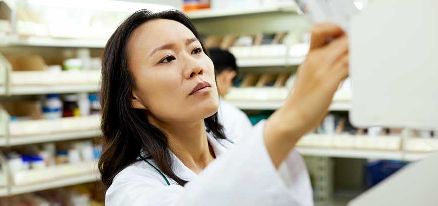 A pharmacist reaches high on a shelf for a box