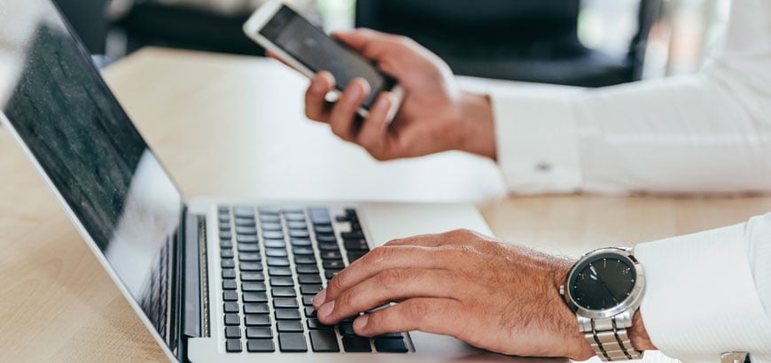 Close up of hands holding a mobile phone and typing on a computer.