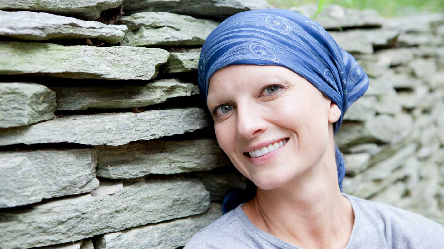 A cancer patient in a head scarf smiles in front of a brick wall