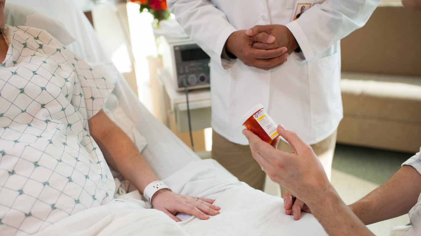 A patient laying in a hospital bed, a person standing next to her and a healthcare worker showing her a pill bottle.