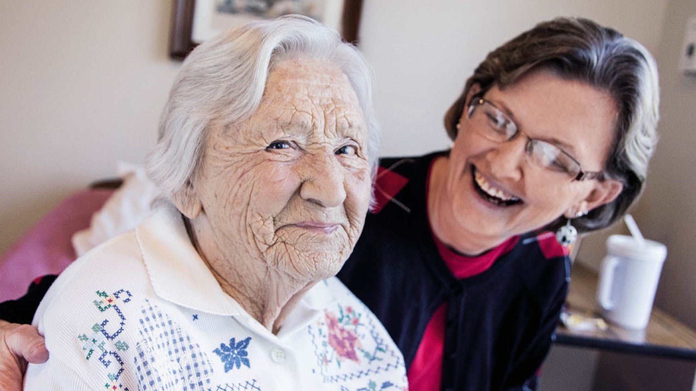 A woman poses and smiles with an elderly woman inside a care home