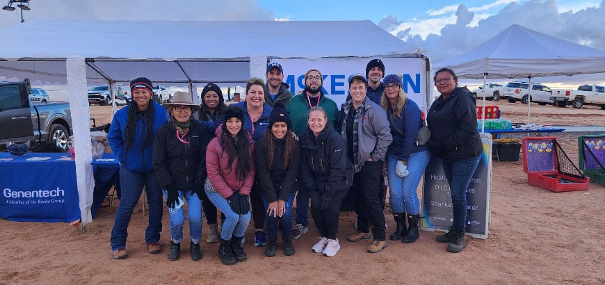 A group of people pose in front of a McKesson banner and Genentech table under a tent, with parked trucks and a cloudy sky in the background.