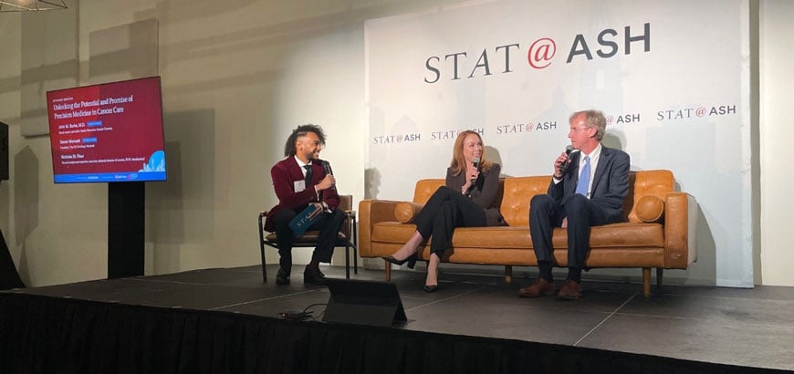 Three people sit on a stage discussing under a "STAT@ASH" sign, with a presentation screen nearby.