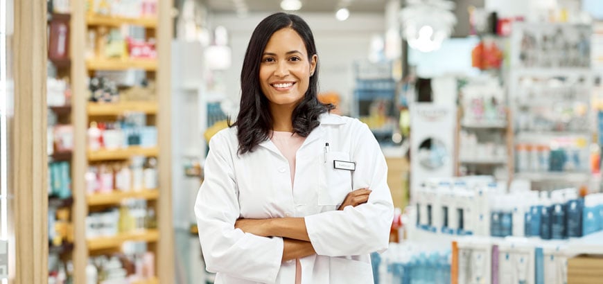 A pharmacist in a white coat stands with arms crossed in a pharmacy aisle.