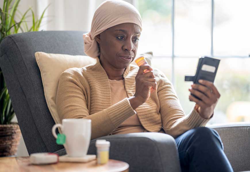 A woman holds her cell phone in one hand and her pill bottle in the other while she relaxes at home