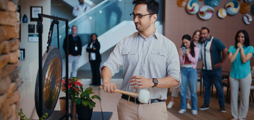 A person strikes a gong in an indoor setting with people watching in the background.