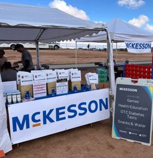 People gather under a tent with McKesson and Genentech banners, featuring health education materials and products, against a backdrop of parked trucks and cloudy sky.