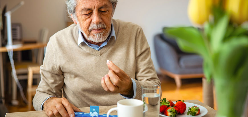 An elderly person sits at a table with a pill, strawberries, and a glass of water.