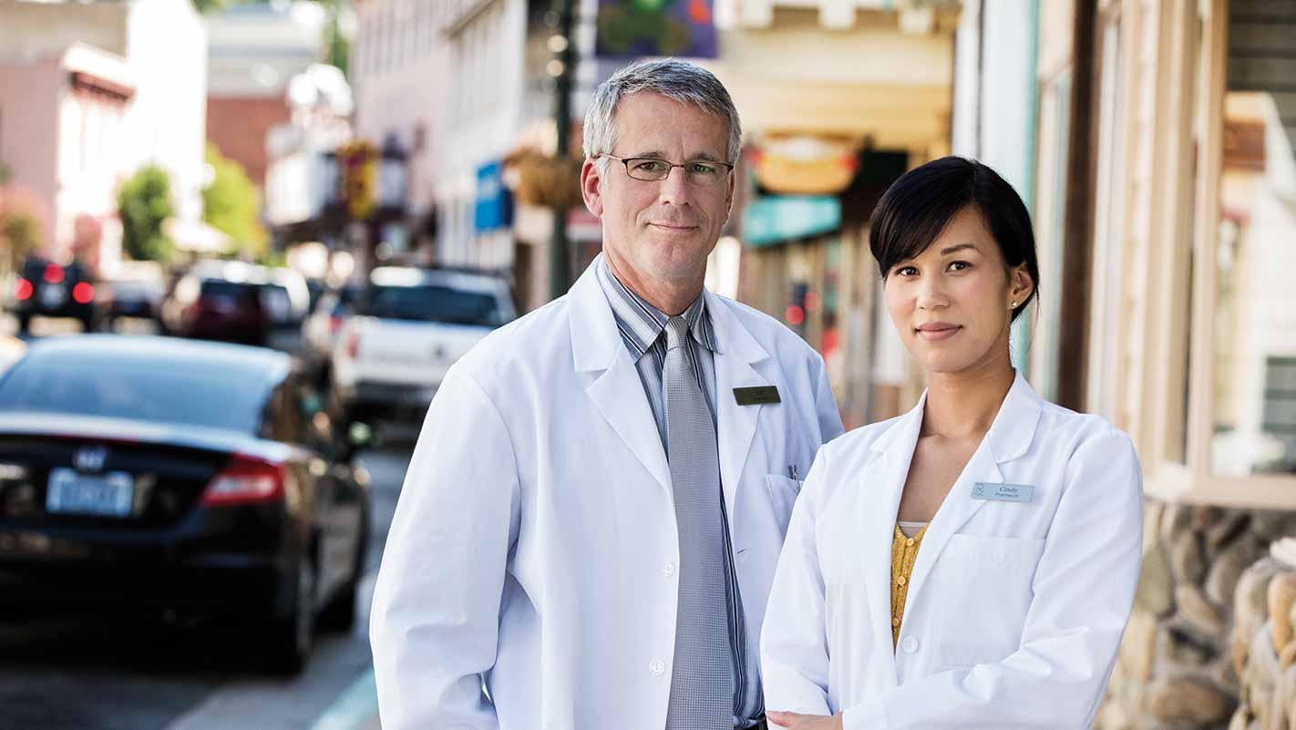 Two people wearing white lab coats stand on a street with blurred faces. The background shows parked cars and buildings.