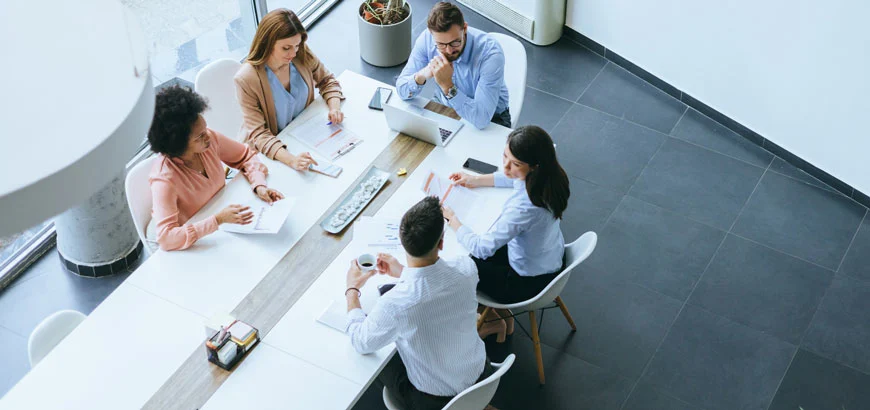 Five people having a meeting around a long table with laptops and documents.