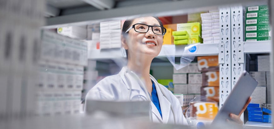 A pharmacist in a white coat holds a tablet while standing in front of shelves filled with medication boxes.