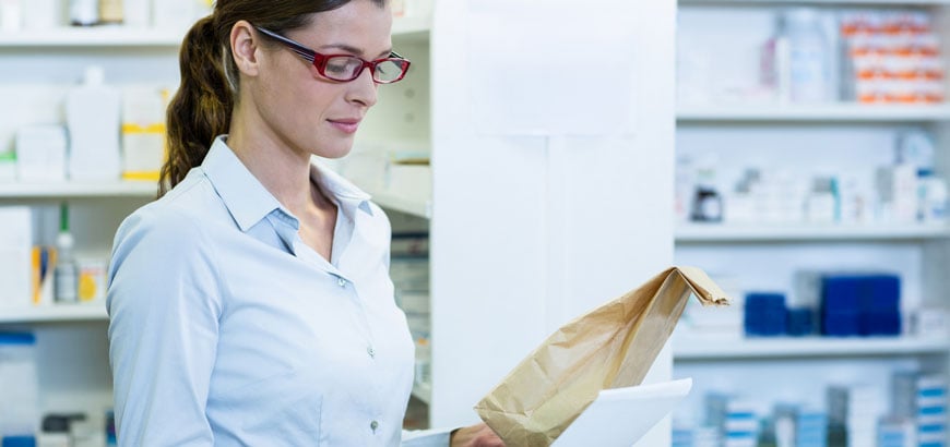 A pharmacist in a white coat holds a brown paper bag and a document in a pharmacy setting.