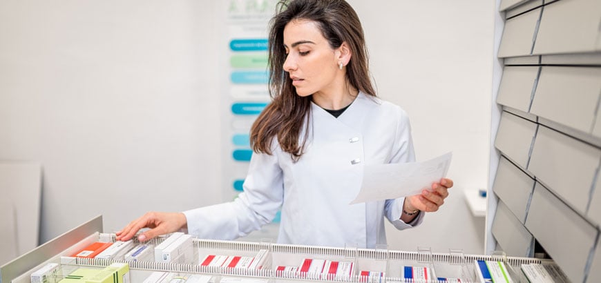 Pharmacist organizing medications in a drawer while holding a sheet of paper.