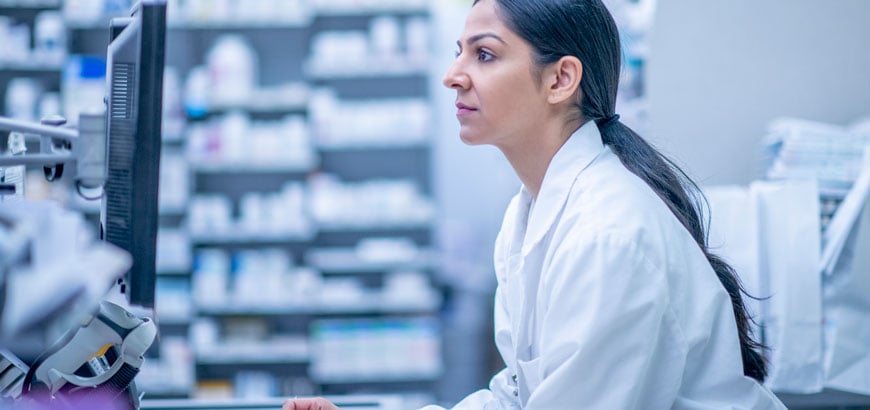 Person in a lab coat working at a computer in a pharmacy with shelves of medications in the background.