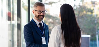 Two professionals are standing and conversing in a well-lit, modern office setting.