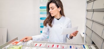 A person in a lab coat organizing medication boxes in a pharmacy, holding a paper.