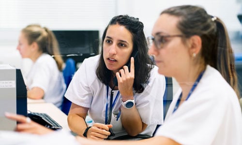 Two healthcare workers sit together at a desk and look at a computer screen.<br>  